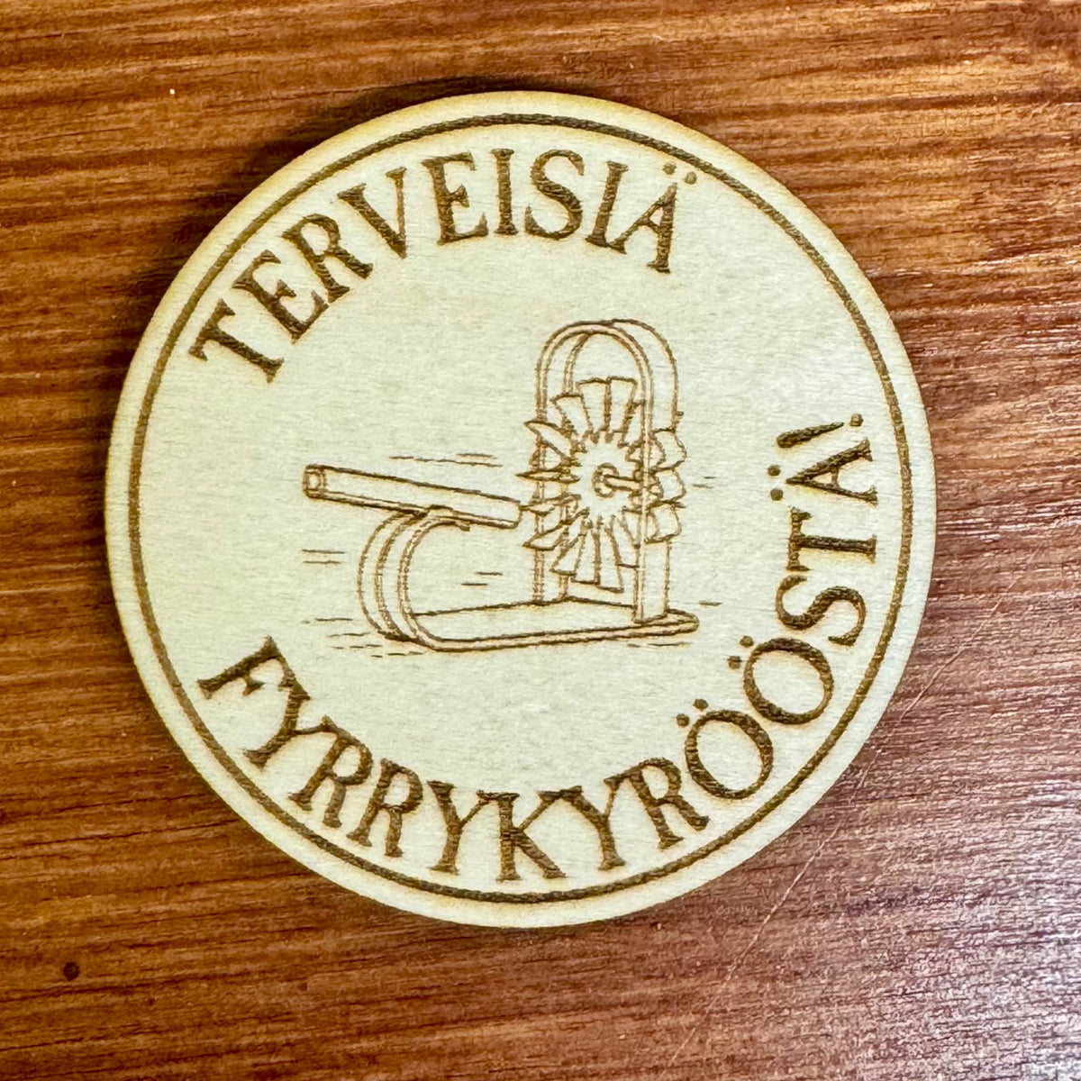 Pyöreä puinen jääkaappimagneetti, jossa on pläkkyri-aihe ja teksti Terveisiä Fyrrykyrööstä!